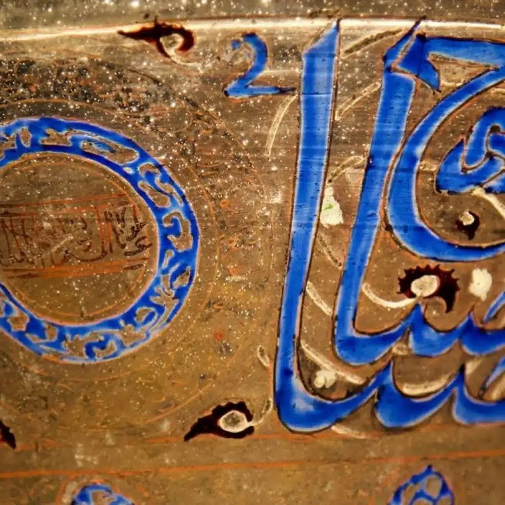 This image shows a close-up of ancient Arabic calligraphy painted on a shiny, glazed ceramic surface. The script is written in bold blue and brown strokes with intricate circular patterns and decorations surrounding the text. The letters are beautifully curved and flow across the surface, highlighting traditional Islamic art and craftsmanship.